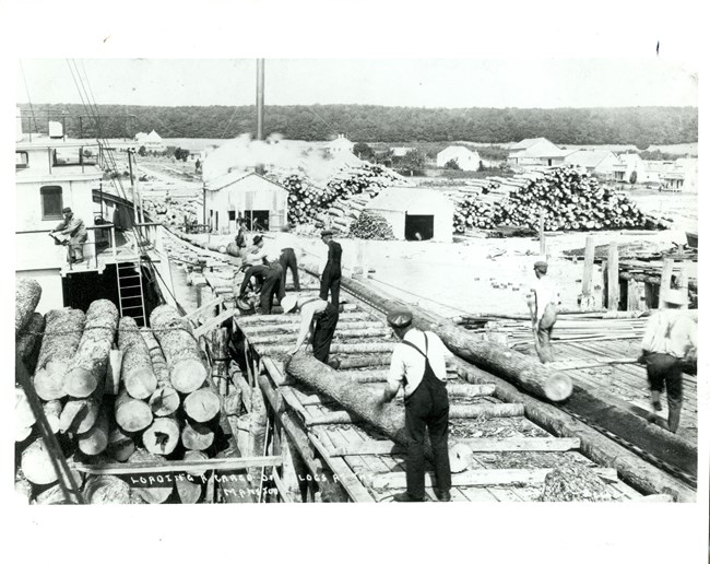 Workers move along a long dock loading cut logs onto a ship. In the background are many buildings and tall stacks of logs. Words along the bottom read: “Loading cargo of logs at the Manitou.”