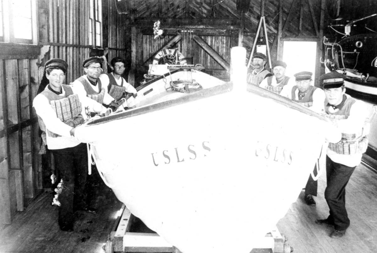 The smiling, heroic crew of the South Manitou Life Saving Station stand around their trusty surfboat, ready to respond to a ship’s distress at a moments notice.