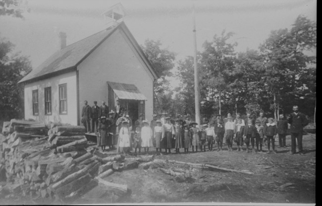 The residents of South Manitou Island stand proudly around their newly constructed schoolhouse. The first class of students await their first lesson as eagerly as most children going to school.