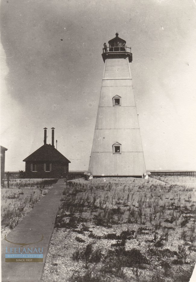 A tall, pillar shaped lighthouse stands on a sandy beach. A white tower with a black top. Behind it sits a shorter fog signal building with two smokestacks protruding from the roof. A vigilant lighthouse keeper stands on the catwalk at the top.