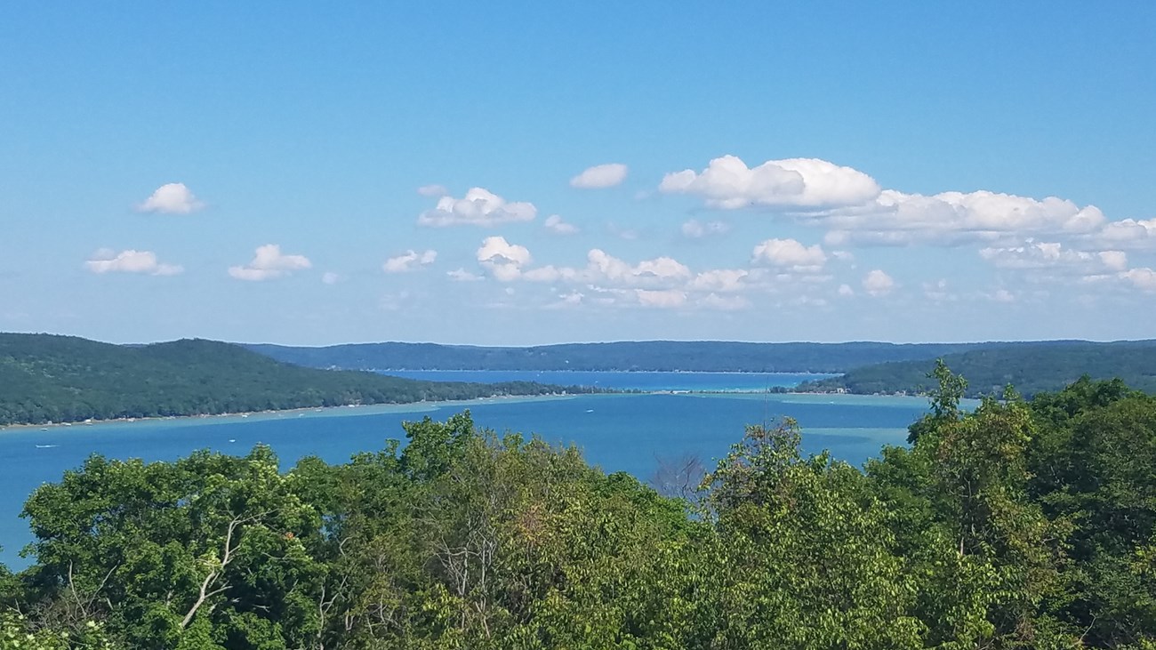 From behind green treetops, bright blue Glen Lake a few fluffy clouds paint the sky.