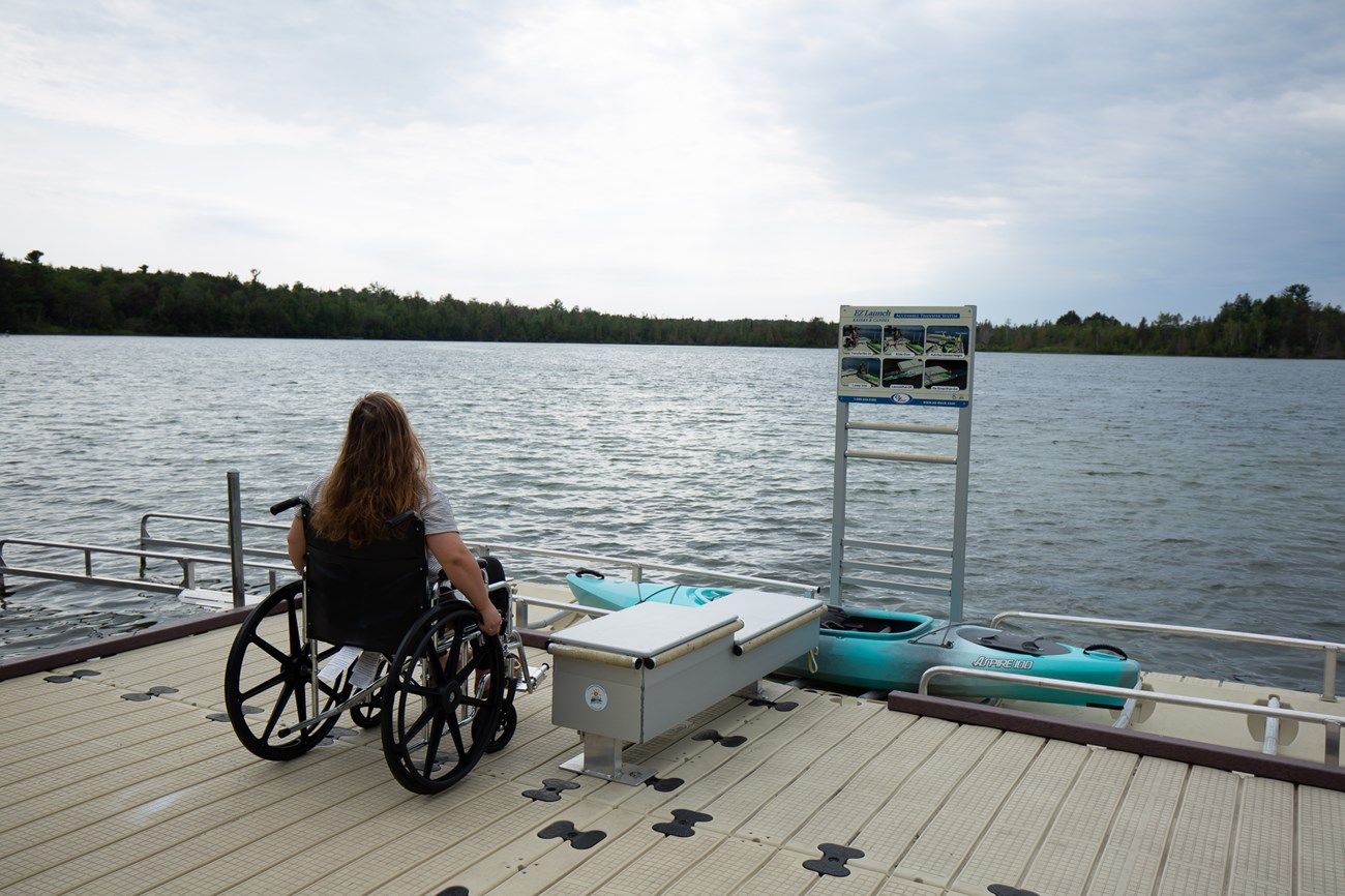 Person sitting in a wheelchair sits next to a step-down seat leading to a kayak nested in a launch with rollers and handrails.