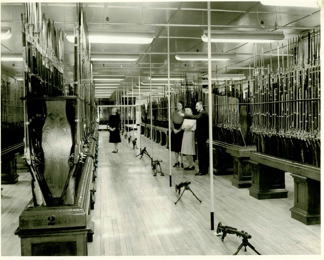 Four people are assessing large rows of muskets that are standing upright on raised tables specially designed to hold guns. A row of bipod equipped machine guns are lined up along the floor.