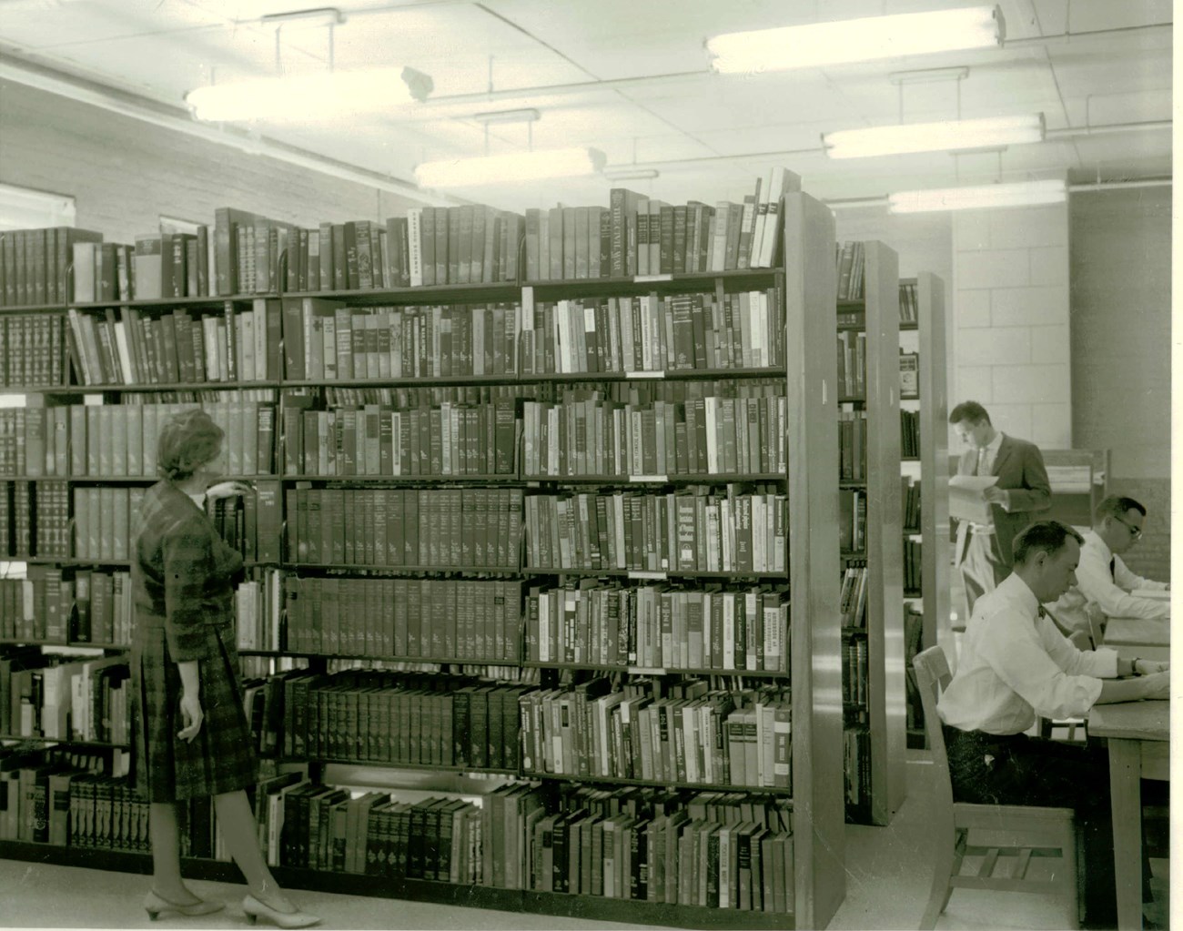 A woman in a dress peruses over a row of books on one of three large library shelves. Two men sit at a desk on the right side and a fourth person looks at a book near the furthest shelf in the back.