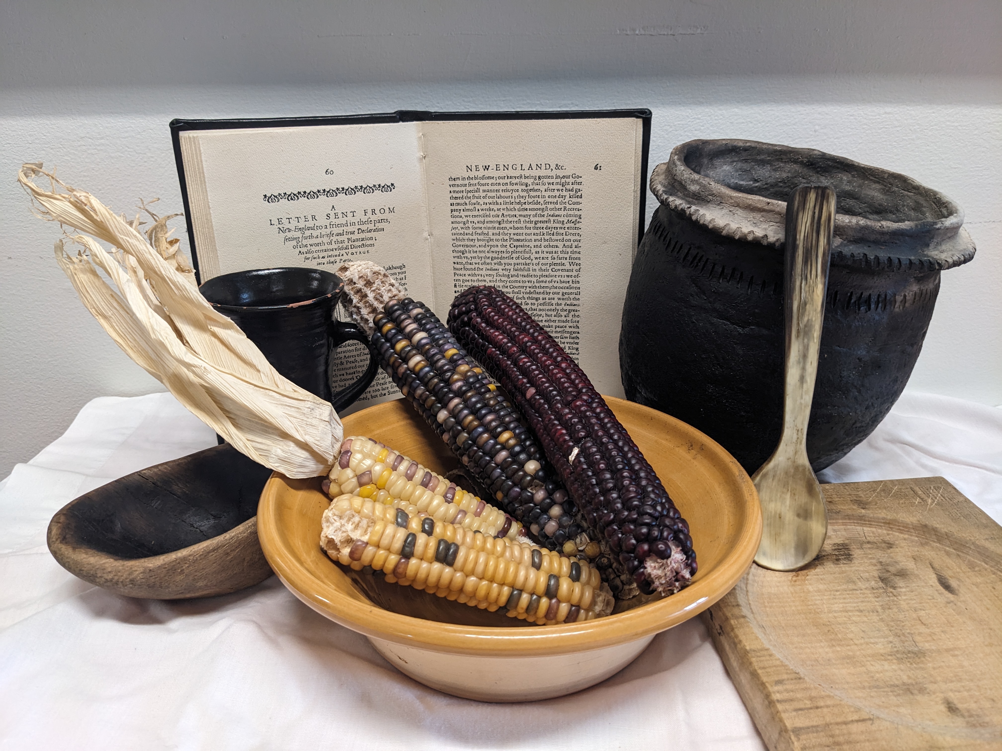 Corn cobs sit in a wooden bowl, surrounded by a book, sppon, bowl and mug.