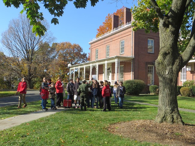 A group of students standing on a walking path outside of a building with a white porch.