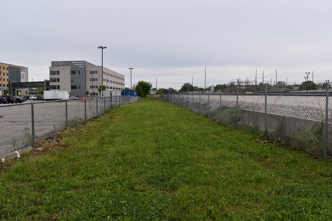 You are looking down a long strip of grass surrounded by a chain link fence. Railroad tracks are on the right. A parking lot and hospital are on the left side in the distance.