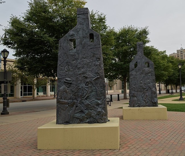 Two metallic appearing monuments stand on a brick walkway in a park. The objects look like silhouetted homes with chimneys. Detailed relief images depicting scenes from the riots are etched into the metal.