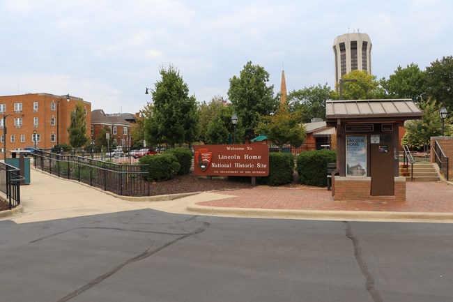 On the left, a paved ramp leads up to a walkway. In the center, there is a brown "Welcome to Lincoln Home National Historic Site" sign and a small brown kiosk. On the right, there is a paved set of stairs.