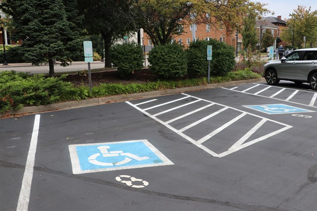 Three accessibility-reserved parking spaces are marked with white lines and blue wheelchair symbols. There are signs in front of each spot to mark them. There is a silver vehicle in one spot.