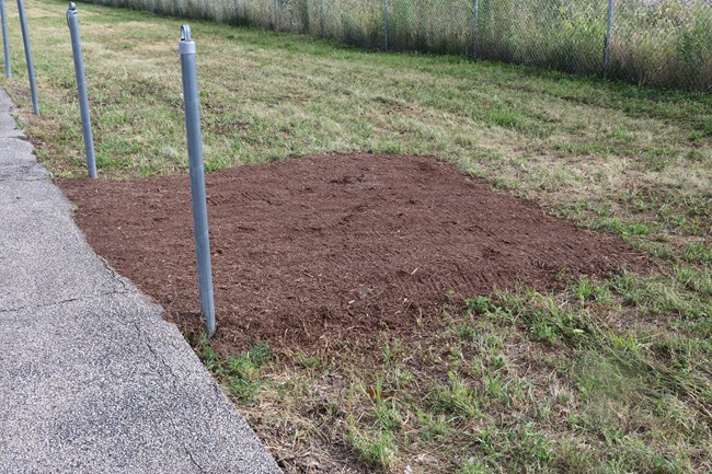Between two metal poles, a brown, mulch ramp leads from a section of pavement to a section of grass.