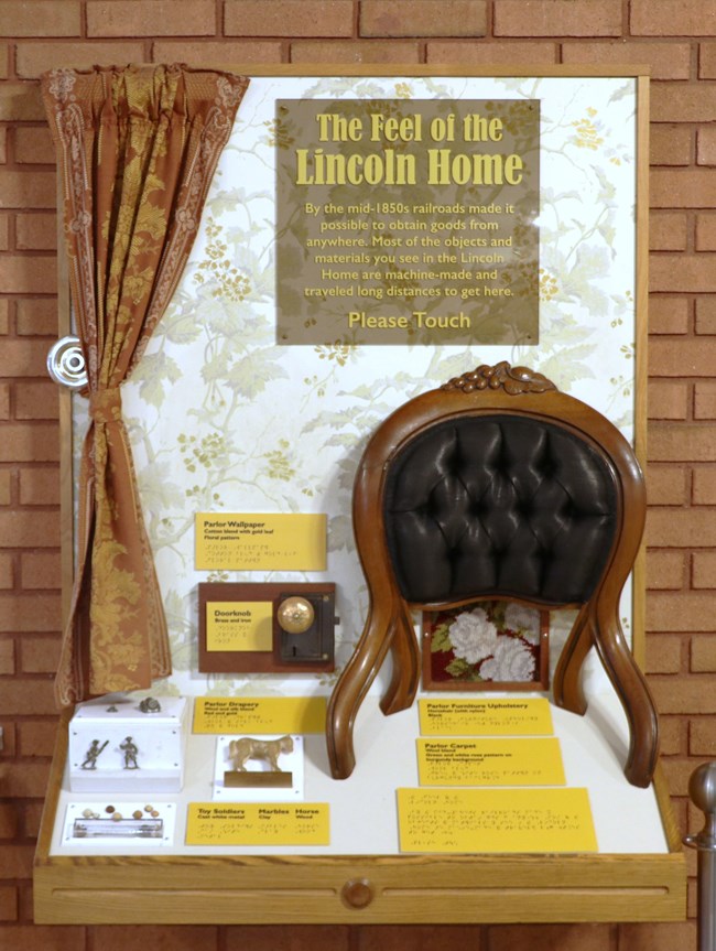 An exhibit titled, "The Feel of the Lincoln Home". There is a curtain, floral wallpaper, metal doorknob, wooden chair with black upholstery, and several toys for visitors to touch.