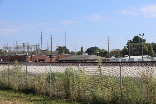 A set of train tracks run across the image with gravel on either side. There are a fence and grass in front of the tracks.