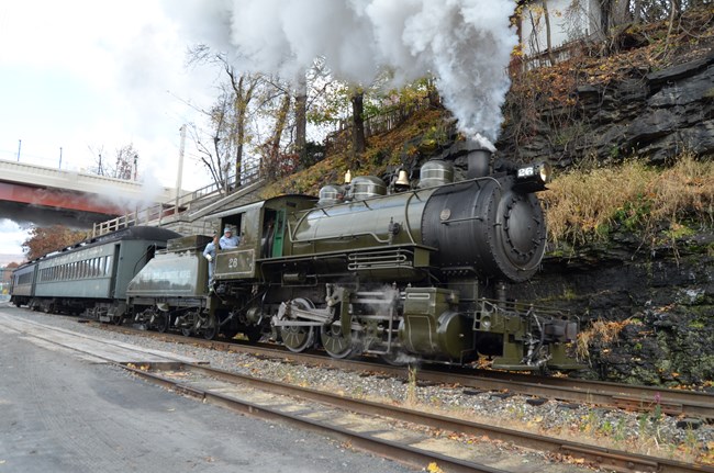 An engineer leans out the cab window of dark green steam locomotive moving towards camera