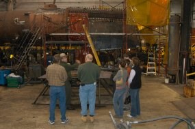 a park ranger tells a small group of visitors about the different projects in the locomotive shop.  behind the group is a stripped down steam locomotive
