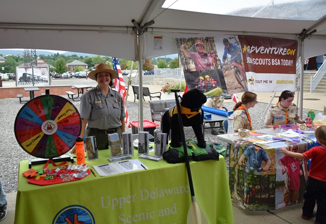 Ranger and Scouts at tables under a tent with brochures, photos, a game wheel and stuffed animal displayed on the tables.