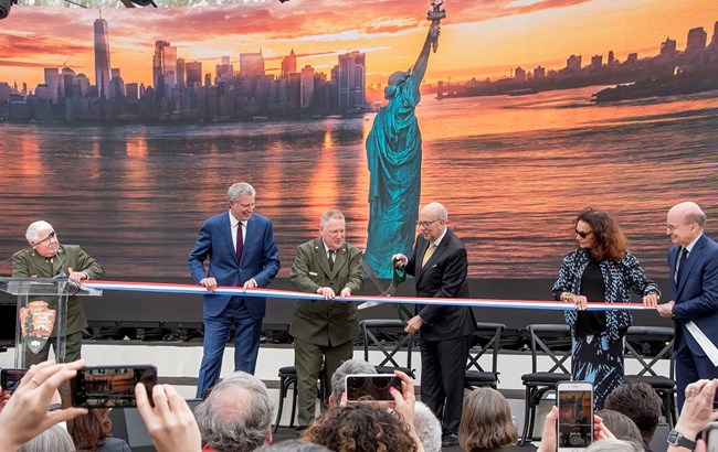 A ceremonial ribbon is cut in front of a large panoramic photo of the Statue of Liberty.