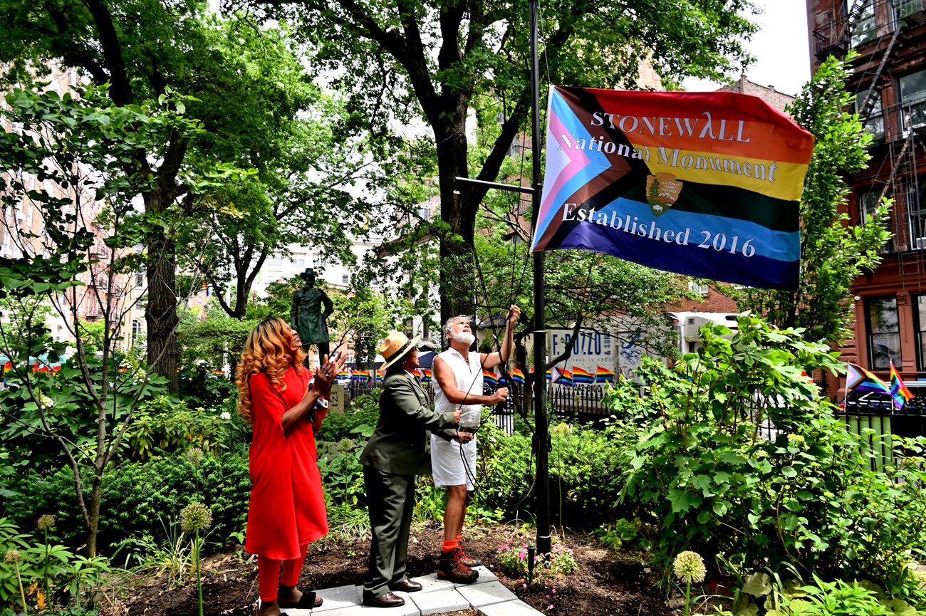 Artist Miss Simone, NPS Superintendent Shirley McKinny, and LGBTQ+ activist Steven Love Menendez raise the Progress flag at Christopher Park.
