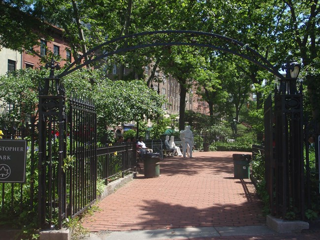 Gated entrance leads to a brick walkway with benches, trees, and several memorials.
