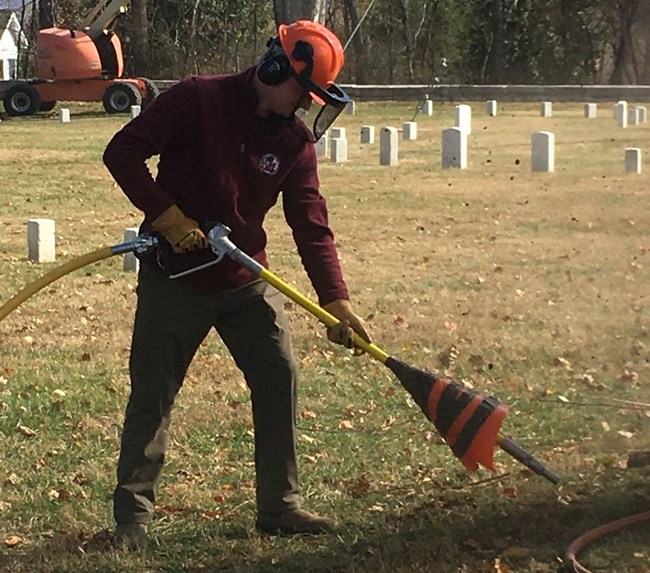 A young man wearing a hard hat and maroon shirt operates an air spade.