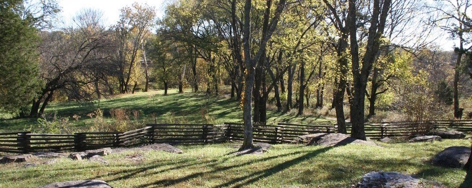 Landscape photo with trees, hills, wooden fence, and green grass and rocks in foreground.