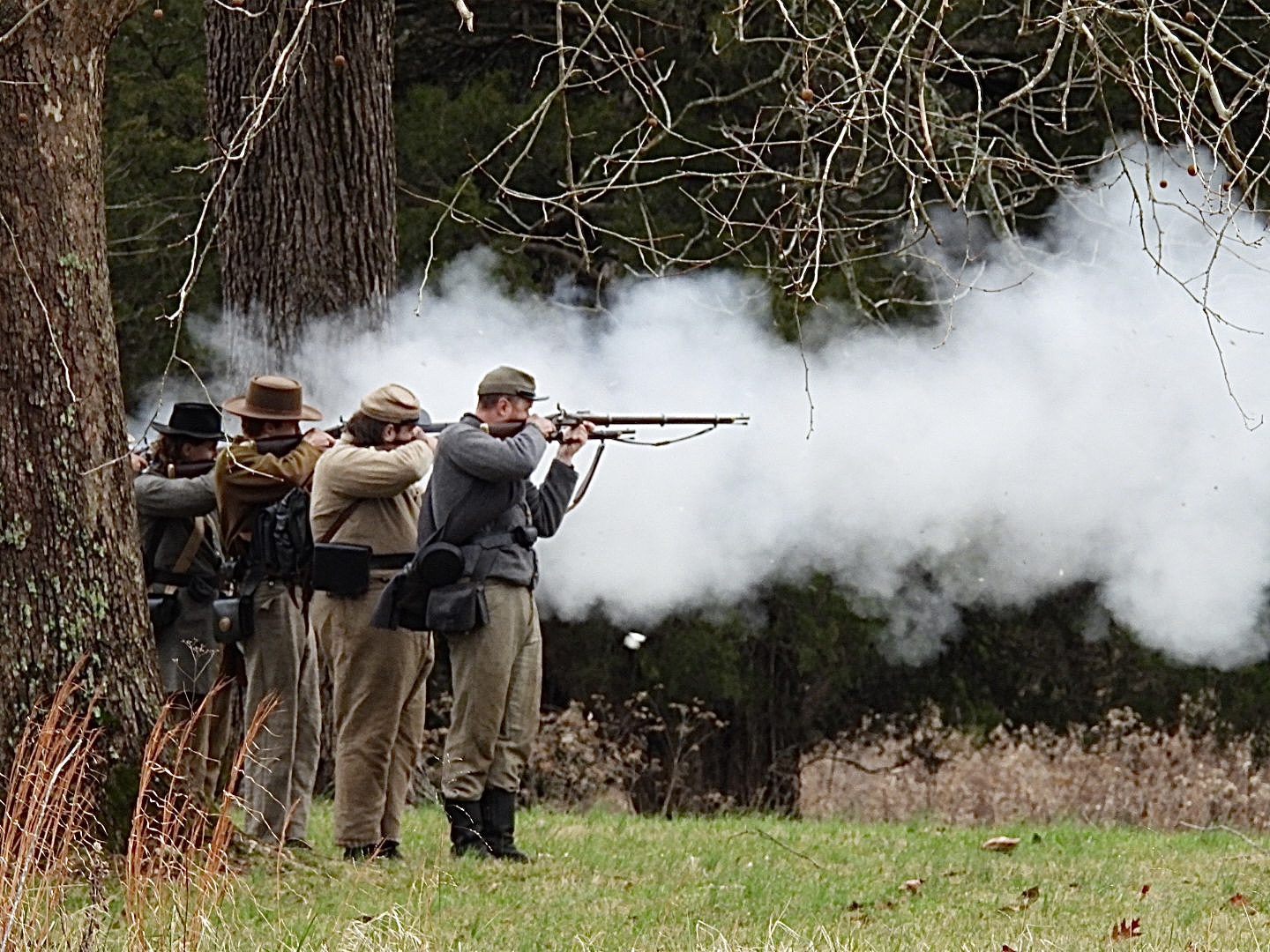 Men in gray uniforms stand in a line firing muskets.