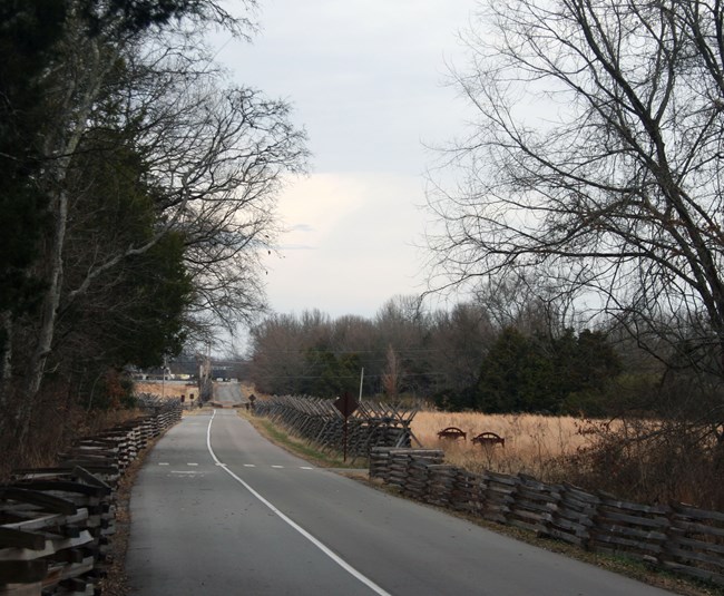 A paved road is bordered by snake rail fence on both sides. Cannon silhouettes stand in a field to the right.