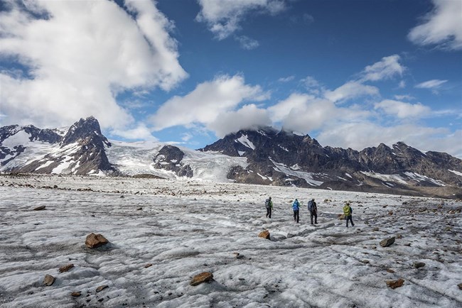 A group of 4 people walk across a glacier with mountains in the background.