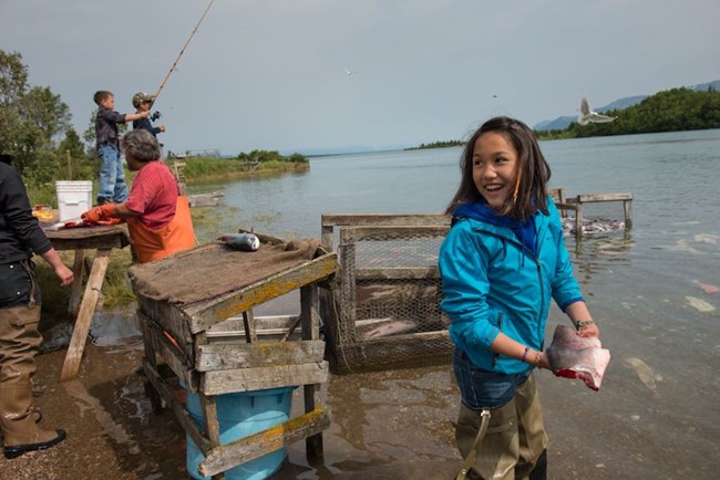 A young girl stands on the edge of water holding a piece of clean, cut fish.