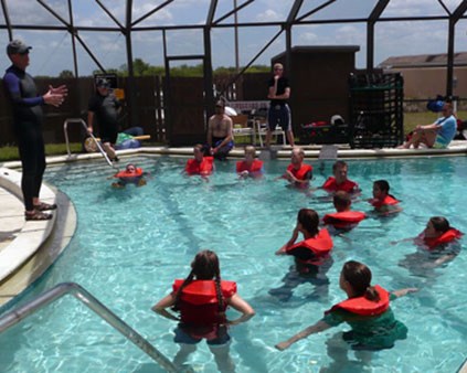 Several people in orange life jackets float in a pool while looking up at a man standing on the edge of pool