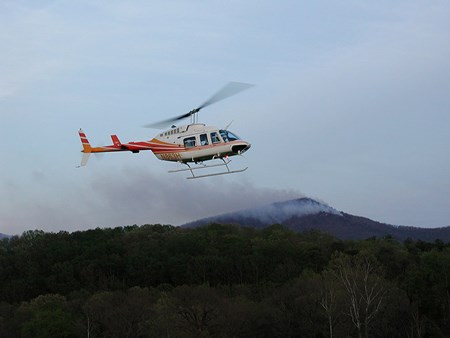 A helicopter hovers over trees while smoke drifts in the background