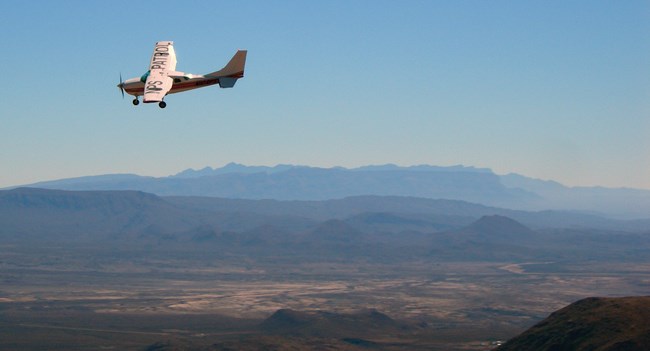 A plane flies high over a desert landscape