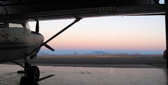 A plane sits in a dark hanger with a view of the sunset over the mountain range