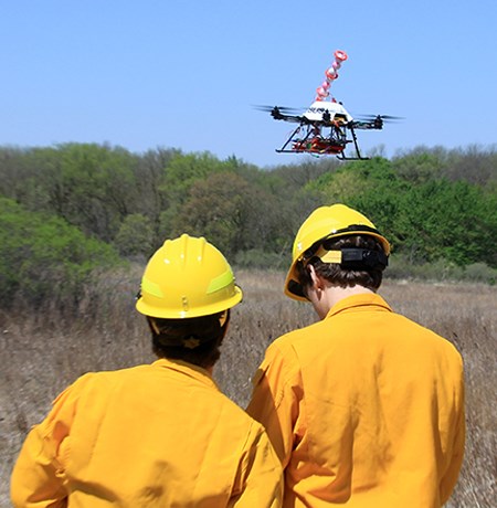 Two people in yellow helmets and shirts operate a drone aircraft above dry vegetation