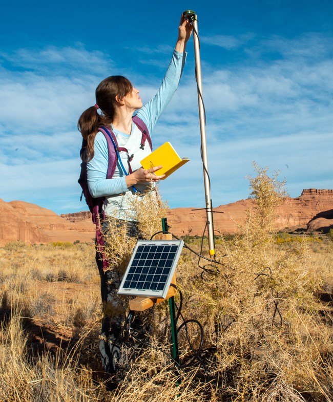 a woman sets up acoustic monitoring equipment powered by a small solar panel in the field in a desert landscape