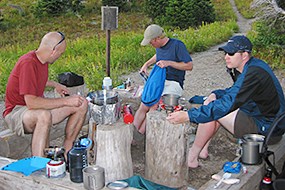 Three men sit in a backcountry food preparation area cooking food