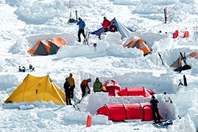 Yellow, orange and red tents are set up in snow with people standing next to them