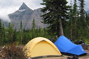 A yellow and blue tent is set up in front of mountains. Rains clouds are present.