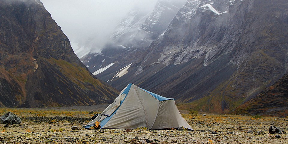 A gray tent is set up in the tundra with snow mountains behind it