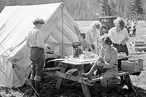 A historic black and white photo of five women camping with period camping gear and a canvas wall tent
