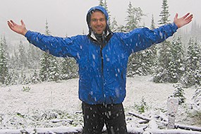 A man in a blue raincoat stands in a summer snowstorm with his arms extended