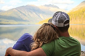 A man wraps his arm around a woman who is resting her head on his shoulder while looking at mountains