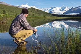 A man kneels to filter water while looking at snow-capped mountains
