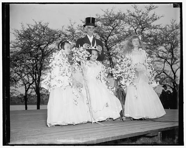 Three girls in formal white dresses adorned with cherry blossoms stand on a stage with a man in a top hat. The man is placing a crown on the head of the girl in the middle.