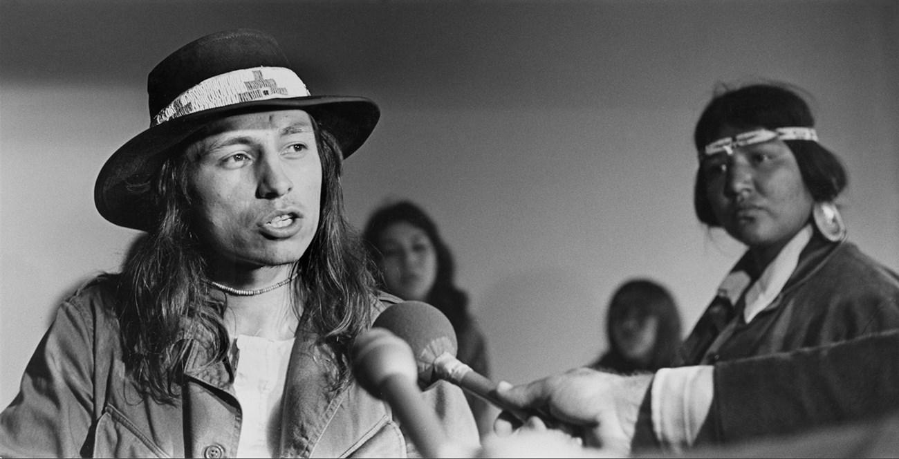A young male wearing broad-brimmed hat with beaded band stands before two microphones