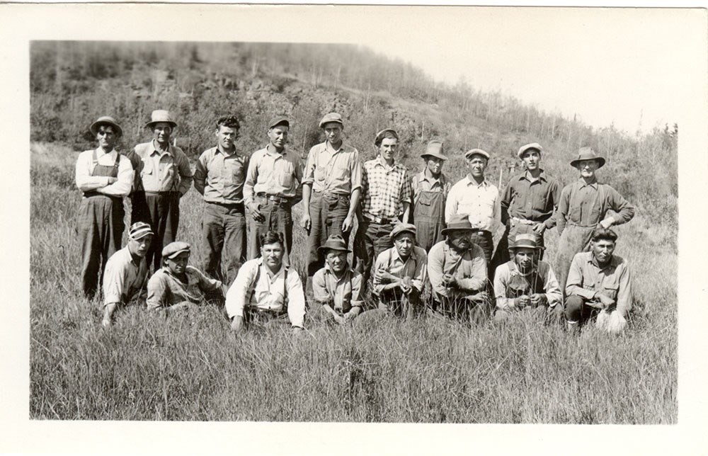 Historic photo of two rows of men in work clothes, a row on the grassy ground in front of a standing row