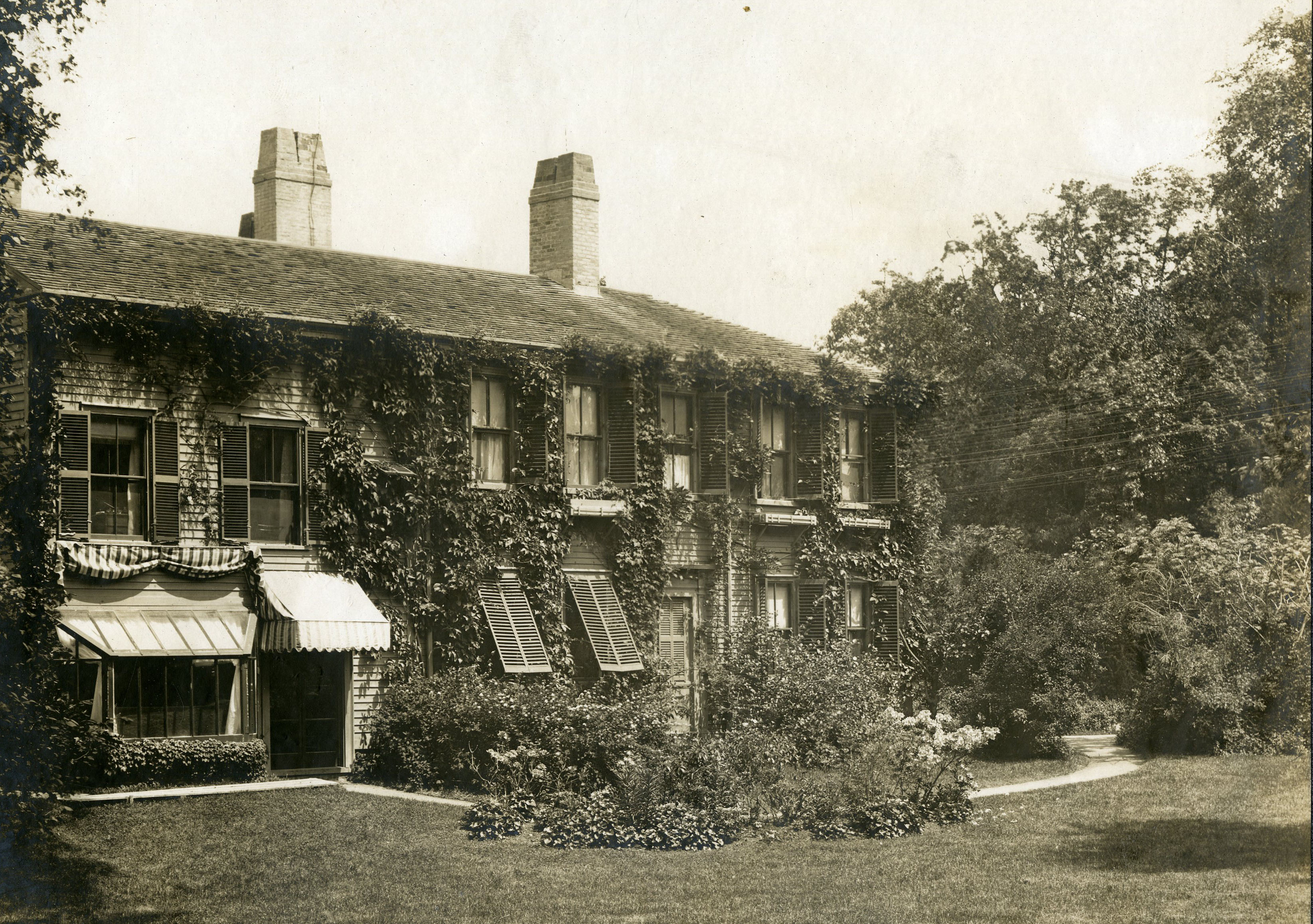 Ivy grows around the windows, with shutters and awnings, of a two-story house with siding.