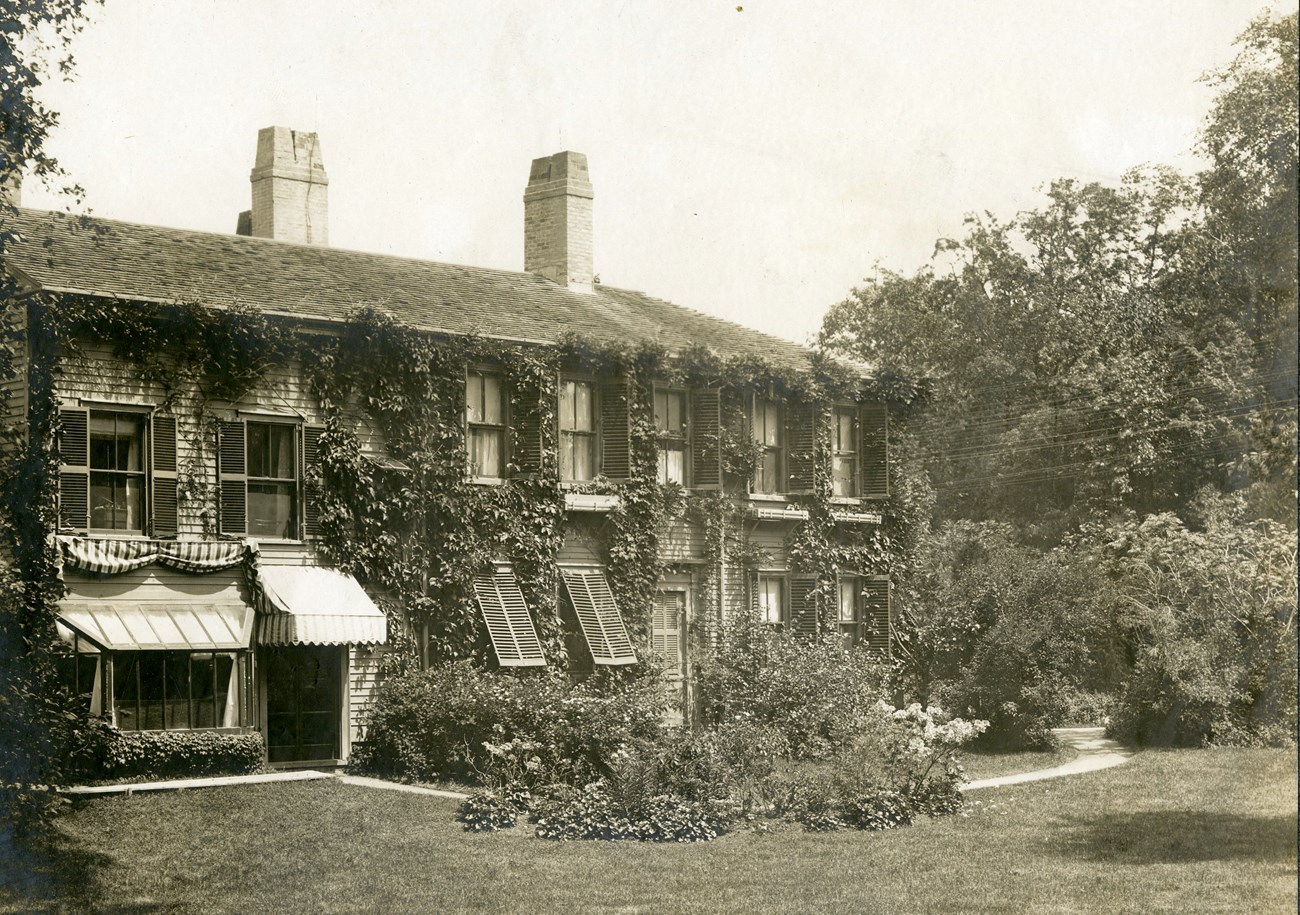 Ivy grows around the windows, with shutters and awnings, of a two-story house with siding.