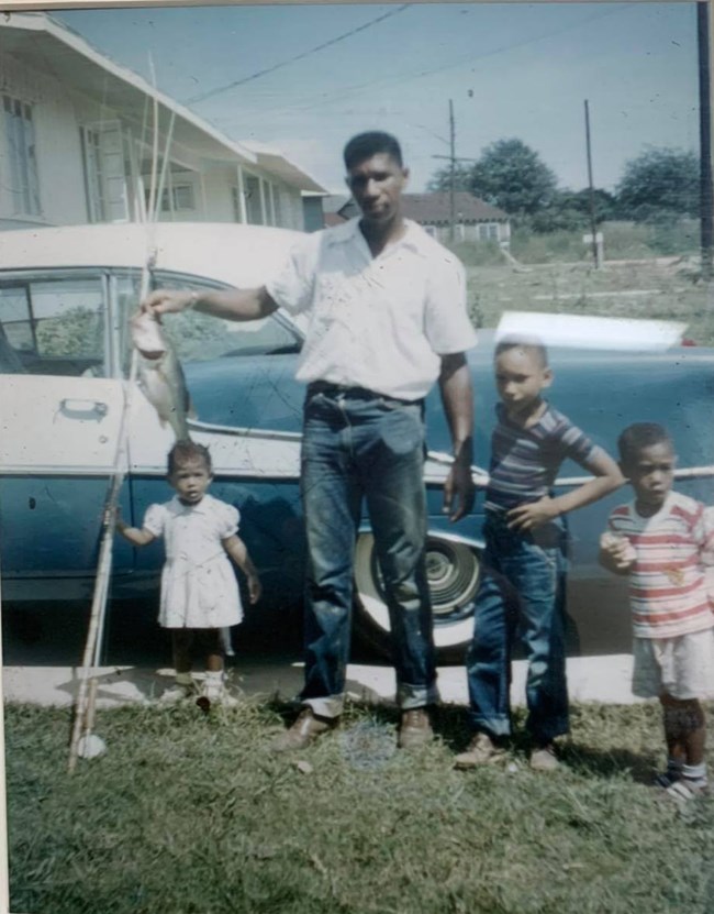 Medgar Evers holding a fish next to three young children, in front of a 1950's blue and white car in a driveway.