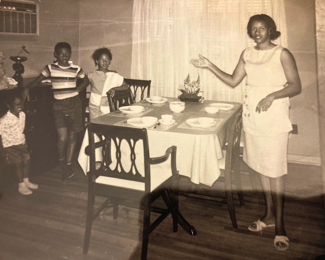 Myrlie Evers stands beside a table set with dishes and silverware, one arm outstretched. Three children stand on the opposide side of the table. Long, sheer curtains cover the window. (sepia-toned)